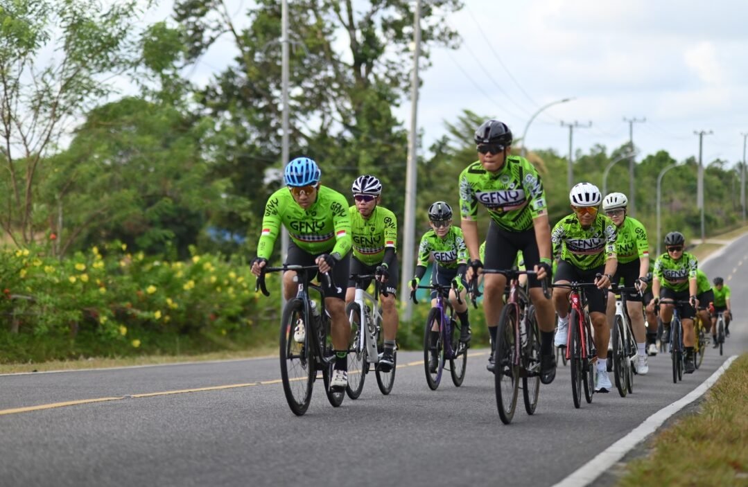Rombongan pesepeda mengenakan jersey hijau GFNY melintasi jalan raya aspal yang mulus dikelilingi pepohonan hijau saat mengikuti kegiatan Group Ride Road to GFNY Belitung 2026. (Dok. HO/Faktakalbar.id)