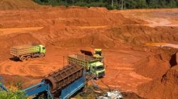 Foto sebuah area pertambangan di Simpang Dua, Kabupaten Ketapang. Terlihat lahan luas dengan tanah berwarna merah, truk pengangkut material, serta alat berat yang digunakan dalam aktivitas penambangan. Vegetasi di sekitar lokasi tampak berkurang, mengindikasikan dampak dari eksploitasi lahan.