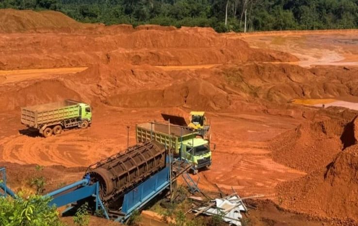 Foto sebuah area pertambangan di Simpang Dua, Kabupaten Ketapang. Terlihat lahan luas dengan tanah berwarna merah, truk pengangkut material, serta alat berat yang digunakan dalam aktivitas penambangan. Vegetasi di sekitar lokasi tampak berkurang, mengindikasikan dampak dari eksploitasi lahan.