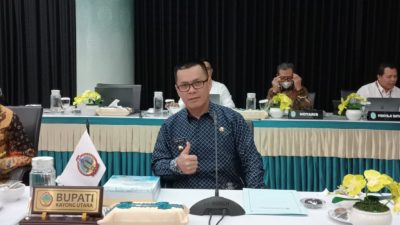 Bupati Kayong Utara, Romi Wijaya berkemeja batik biru duduk di meja rapat dengan papan nama "Bupati Kayong Utara" di depannya. Ia memberikan gestur jempol ke arah kamera. Di sekelilingnya, beberapa peserta rapat lainnya terlihat sibuk dengan dokumen dan perangkat kerja. Latar belakang menunjukkan ruang rapat dengan dekorasi hijau dan hitam serta meja yang dihiasi taplak berwarna hijau tosca dan krem.