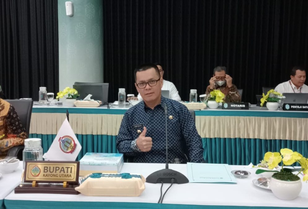 Bupati Kayong Utara, Romi Wijaya berkemeja batik biru duduk di meja rapat dengan papan nama "Bupati Kayong Utara" di depannya. Ia memberikan gestur jempol ke arah kamera. Di sekelilingnya, beberapa peserta rapat lainnya terlihat sibuk dengan dokumen dan perangkat kerja. Latar belakang menunjukkan ruang rapat dengan dekorasi hijau dan hitam serta meja yang dihiasi taplak berwarna hijau tosca dan krem.