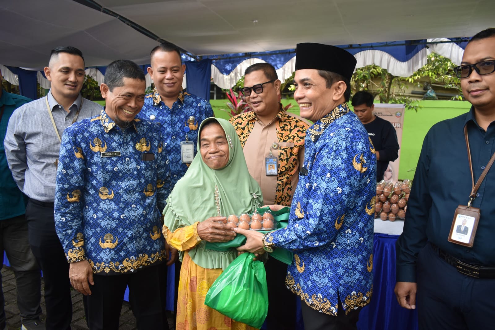 Seorang perempuan lanjut usia mengenakan hijab hijau menerima paket sembako berisi telur dan bahan pokok lainnya dari Wakil Wali Kota Pontianak Bahasan, yang mengenakan batik biru dengan motif khas. Di sekeliling mereka, beberapa pejabat Pemkot Pontianak tersenyum, sementara latar belakang menunjukkan stan penjualan telur dan spanduk operasi pasar murah.