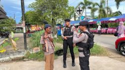 Dua anggota polisi berseragam taktis berbincang dengan seorang pria di pinggir jalan dekat area pasar. Salah satu polisi berkomunikasi menggunakan alat komunikasi, sementara pedagang sekitar beraktivitas di latar belakang.