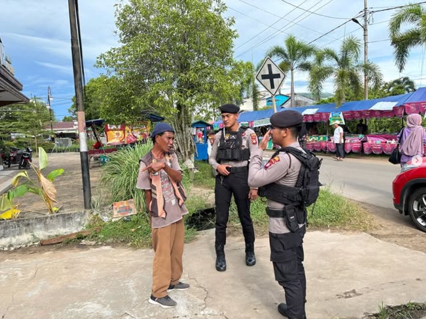 Dua anggota polisi berseragam taktis berbincang dengan seorang pria di pinggir jalan dekat area pasar. Salah satu polisi berkomunikasi menggunakan alat komunikasi, sementara pedagang sekitar beraktivitas di latar belakang.