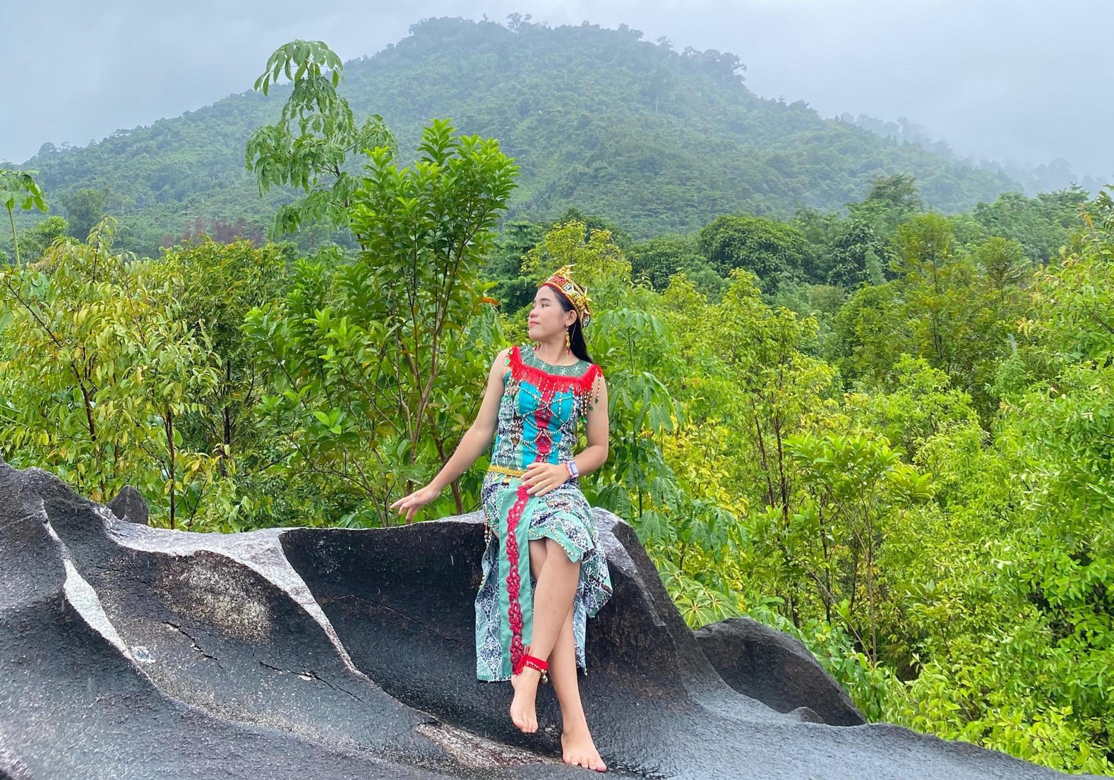 Seorang wanita mengenakan kostum adat Dayak berwarna-warni duduk di atas batu besar di Wisata Batu Kuran, Kecamatan Sajingan Besar, Sambas. Latar belakang menunjukkan hamparan hutan hijau dan pegunungan yang diselimuti kabut tipis.