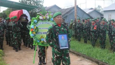 Seorang prajurit TNI mengenakan seragam loreng hijau membawa foto almarhum Sertu Arbuansyah dalam prosesi pemakaman militer. Di belakangnya, rekan-rekan prajurit membawa karangan bunga dan peti jenazah yang ditutupi bendera merah putih, dengan barisan tentara berdiri di sisi jalan