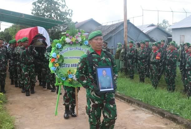 Seorang prajurit TNI mengenakan seragam loreng hijau membawa foto almarhum Sertu Arbuansyah dalam prosesi pemakaman militer. Di belakangnya, rekan-rekan prajurit membawa karangan bunga dan peti jenazah yang ditutupi bendera merah putih, dengan barisan tentara berdiri di sisi jalan