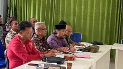 Ade Iswadi sedang berbicara dalam forum diskusi, didampingi peserta lain yang duduk di meja panjang dengan latar belakang tirai hijau.