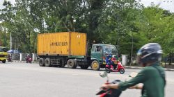 Truk kontainer berwarna kuning dengan tulisan "SAMUDERA" melaju di jalan, dikelilingi oleh pengendara sepeda motor di Pontianak, dengan latar belakang pepohonan hijau dan pagar hitam.