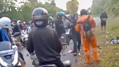 Sekelompok orang berkumpul di jalan dengan beberapa pengendara motor, sementara seorang petugas berseragam oranye berada di sisi jalan. Terlihat sepeda motor dan barang-barang berserakan di lokasi kecelakaan.