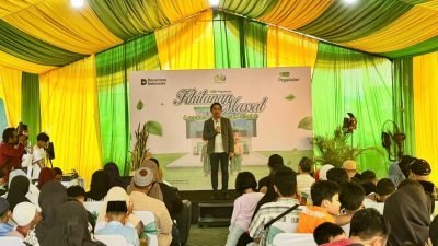 Kadiv Usaha Syariah Holilur Rohman memberikan sambutan di pitanan Massal Pegadaian dengan latar belakang spanduk 'Khitanan Massal Langkah Emas Anak Sholeh'. Terlihat peserta dan orang tua memenuhi tenda acara berwarna hijau kuning