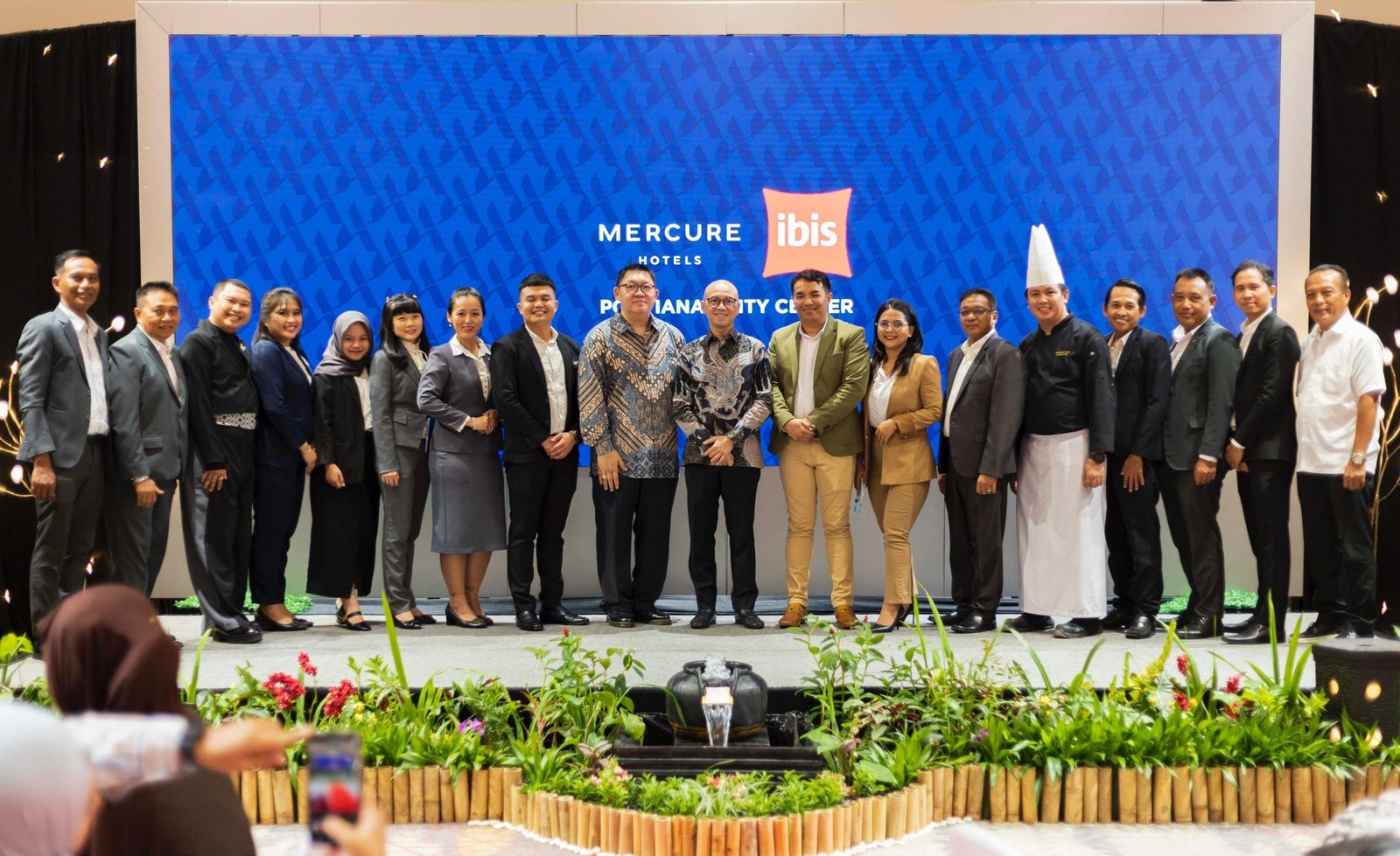 "Kelompok besar staf hotel Mercure dan ibis Pontianak City Center berpose di panggung dengan latar belakang layar besar berlogo hotel. Di tengah terlihat General Manager Ulysses Jeffrey dan Director of Operations Accor Indonesia Endrian Hananto."