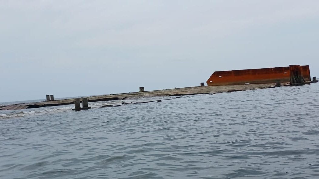 "Bangkai kapal tongkang berwarna oranye dan abu-abu, BG Bukit Emas 2312, terlihat sebagian tenggelam dan kandas di perairan laut yang tenang dengan langit mendung."