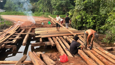 Sekelompok warga laki-laki sedang menyusun papan kayu untuk memperbaiki jembatan rusak di atas sungai keruh di kawasan pedesaan. (Dok. Ist)