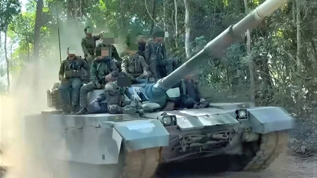 Sejumlah tentara berseragam militer lengkap dengan helm taktis sedang menaiki bagian atas tank tempur utama yang melaju di jalan tanah berdebu di kawasan hutan. (Dok. Ist)