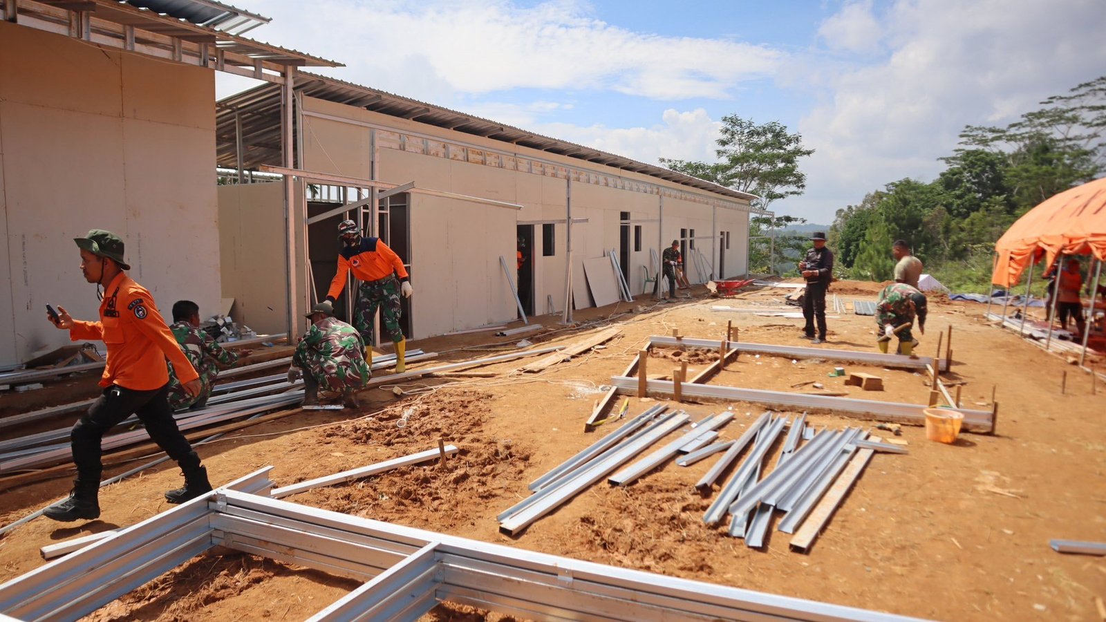 Sejumlah personel TNI dan petugas berbaju oranye sedang bekerja merakit rangka baja ringan untuk pembangunan hunian sementara (huntara) di area tanah lapang yang sedang dikerjakan. (Dok. HO/Faktakalbar.id)