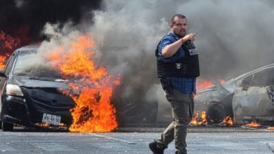 Seorang petugas keamanan berdiri di tengah jalan raya dengan latar belakang dua mobil yang hangus terbakar dan asap hitam pekat mengepul di Meksiko. (Dok. Ist)