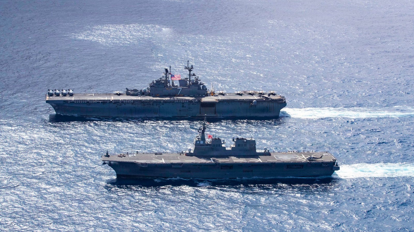 Dua kapal perang militer berlayar berdampingan di lautan lepas, salah satunya adalah kapal serbu amfibi Angkatan Laut Amerika Serikat USS Tripoli (LHA-7). (Dok. DVIDS)