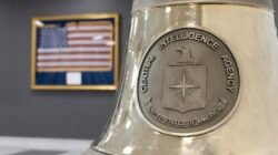 Lencana perak Central Intelligence Agency (CIA) Amerika Serikat yang terukir pada sebuah lonceng besar dengan latar belakang bendera AS. (Dok. HO/Faktakalbar.id)