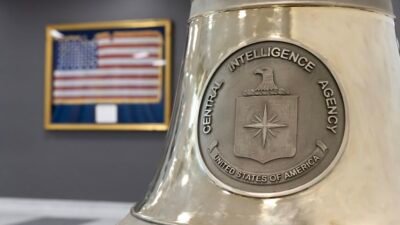 Lencana perak Central Intelligence Agency (CIA) Amerika Serikat yang terukir pada sebuah lonceng besar dengan latar belakang bendera AS. (Dok. HO/Faktakalbar.id)