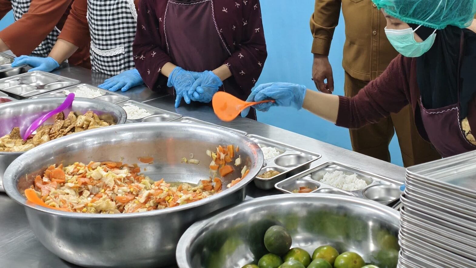 Ilustrasi - Pejabat pemerintah, termasuk gubernur dan bupati yang mengenakan pakaian dinas, sedang meninjau aktivitas pekerja dapur berseragam celemek, masker, dan penutup kepala saat menyiapkan porsi makanan ke dalam baki baja nirkarat. (Dok. HO/Faktakalbar.id)